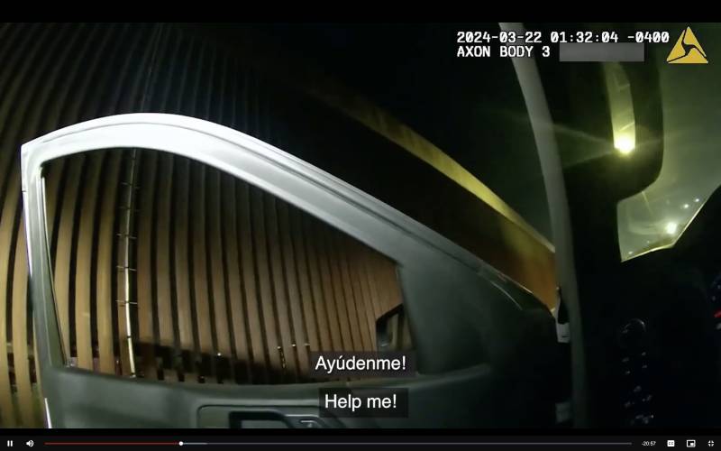 A screenshot from a body-worn camera showing the U.S.-Mexico border wall, with the caption: 'Ayudenme!' or 'Help me!'